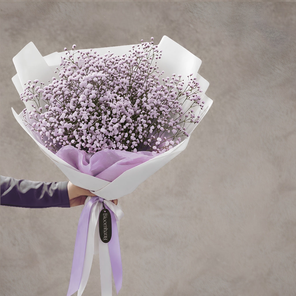 A bouquet of lilac baby's breath flowers wrapped in white paper and a lilaccribbon, held by a hand against a textured gray background.