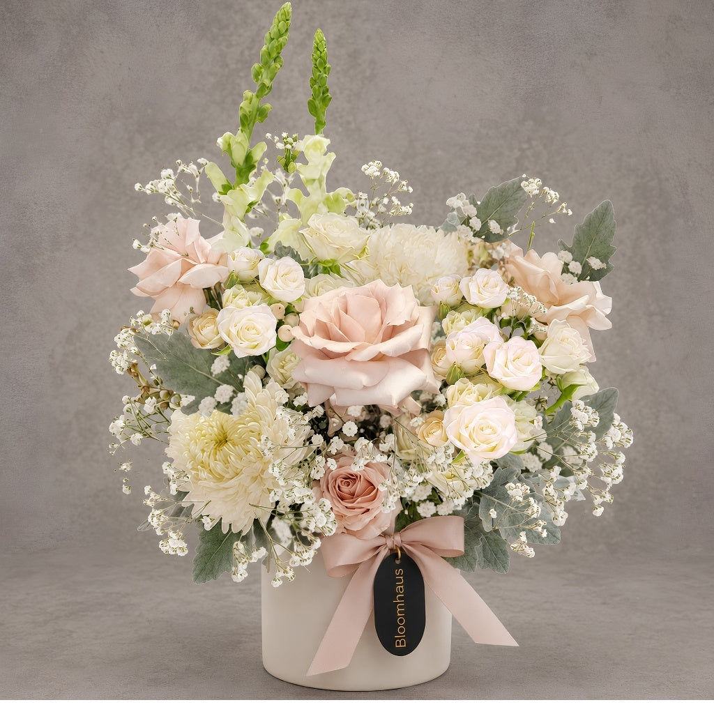 A floral arrangement in a round white box with a pale pink ribbon and a black Bloomhouse tag, photographed against a neutral gray studio background.