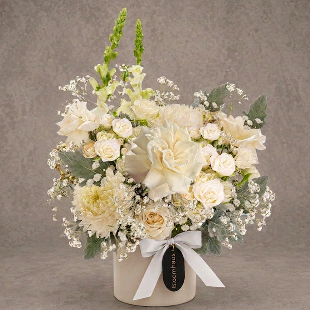 A floral arrangement in a round cream-colored pot, featuring cream and white roses, carnations, baby's breath, and green foliage, tied with a white ribbon and a black Bloomhaus tag, set against a textured gray background.