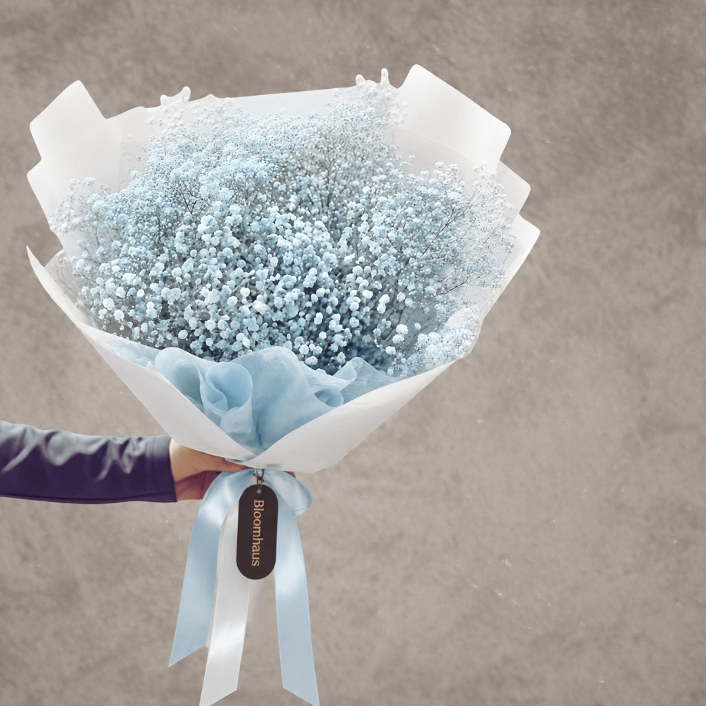 A bouquet of light blue baby's breath flowers wrapped in white paper and a pale blue ribbon, held by a hand against a textured gray background.