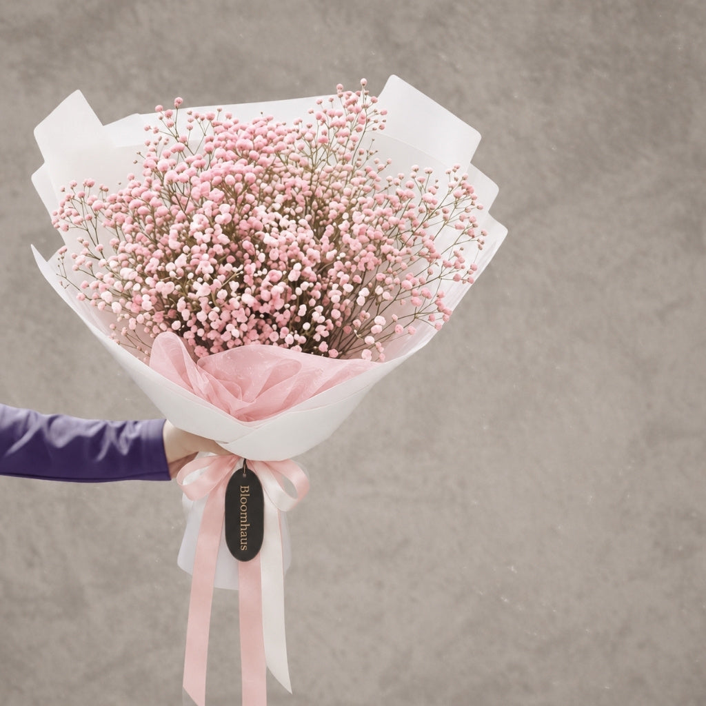 A bouquet of delicate pink and white baby's breath flowers wrapped in white paper with a soft pink ribbon and a black hanging tag, held by a person wearing a purple sleeve against a textured gray background.