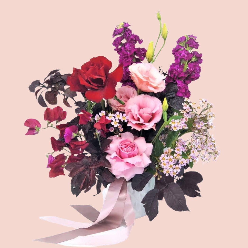 A ceramic pot with an arrangement of red and pink roses and a mix of seasonal blooms, tied with a ribbon.