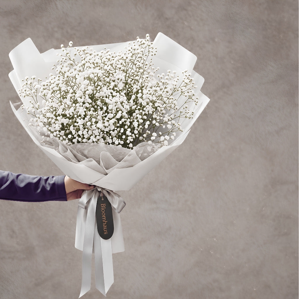 A bouquet of white baby's breath flowers wrapped in white paper and white ribbon, held by a hand against a textured gray background.